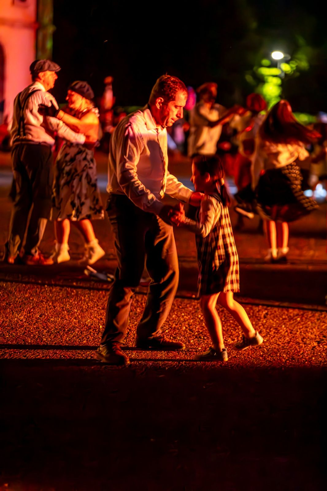 Un homme fait danser une petite fille, au milieu d’autres danseurs en costume d’époque, dans la lumière rouge